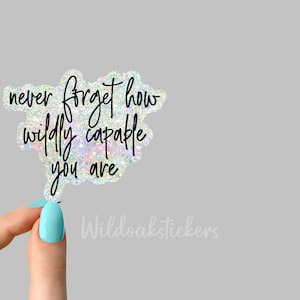 May include: A holographic sticker with the text "never forget how wildly capable you are". The sticker is being held by a hand with light blue nail polish.