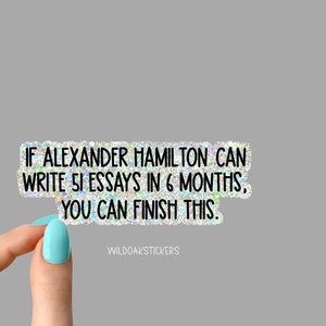 K&ouml;nnte beinhalten: Ein Aufkleber mit glitzerndem, schimmerndem Hintergrund und schwarzem Text, der "IF ALEXANDER HAMILTON CAN WRITE 51 ESSAYS IN 6 MONTHS, YOU CAN FINISH THIS." lautet. Der Aufkleber wird von einer Hand mit hellblauem Nagellack gehalten.