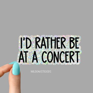 May include: A rectangular sticker with a glittery, iridescent background and black text that reads "I'D RATHER BE AT A CONCERT." The sticker has a scalloped edge and the text "WILDOAKSTICKERS" is at the bottom.