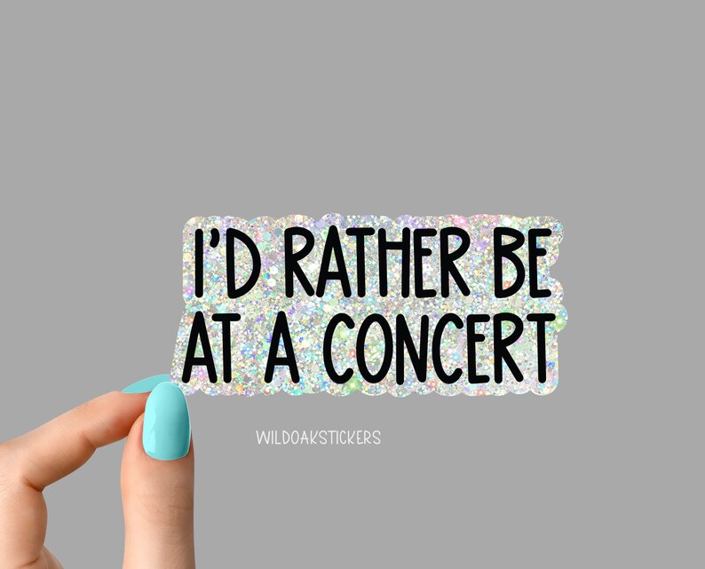 May include: A rectangular sticker with a glittery, iridescent background and black text that reads "I'D RATHER BE AT A CONCERT." The sticker has a scalloped edge and the text "WILDOAKSTICKERS" is at the bottom.