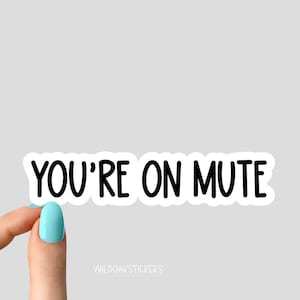May include: White sticker with black text that reads "YOU'RE ON MUTE". A hand is holding the sticker.