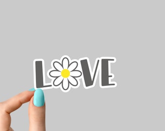 May include: A hand holds up a sticker with the word "LOVE" in black letters with a white daisy in the middle. The daisy has a yellow center.