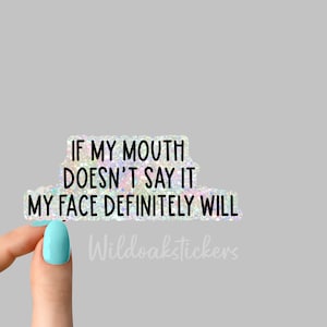 May include: A holographic sticker with the text "If my mouth doesn't say it, my face definitely will." The sticker is held by a hand with light blue nail polish. The text "Wildoakstickers" is visible in the bottom right corner.