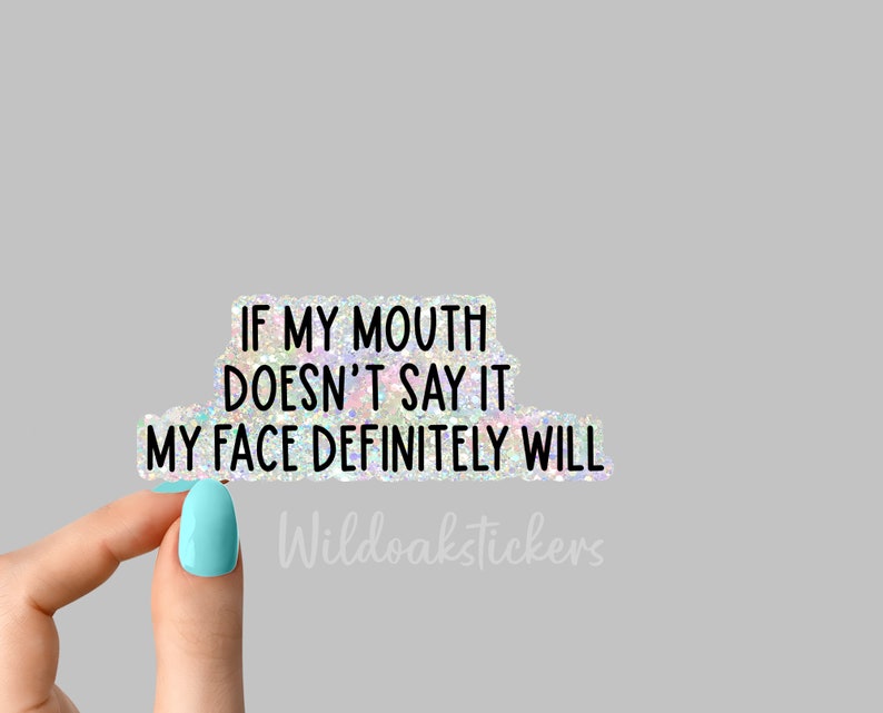May include: A holographic sticker with the text "If my mouth doesn't say it, my face definitely will." The sticker is held by a hand with light blue nail polish. The text "Wildoakstickers" is visible in the bottom right corner.