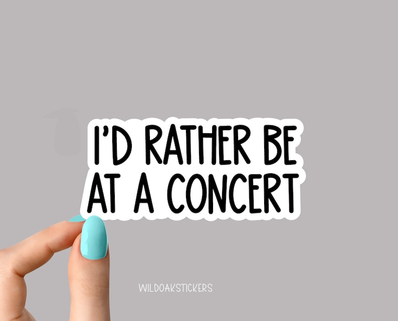 May include: A white sticker with the text "I'D RATHER BE AT A CONCERT" in black, sans-serif font. The sticker is held by a hand with a light blue painted nail. The sticker has a black border. The sticker is on a gray background.