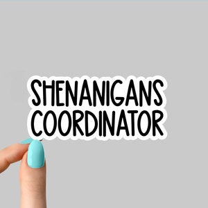 May include: A white sticker with the words "SHENANIGANS COORDINATOR" in a bold, black font. The sticker is held by a hand with a light blue painted fingernail against a gray background.