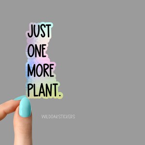 May include: A holographic sticker with the text "JUST ONE MORE PLANT." The sticker is being held by a hand with light blue nail polish.