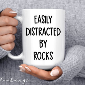 May include: White ceramic coffee mug with black text that reads "Easily Distracted By Rocks".