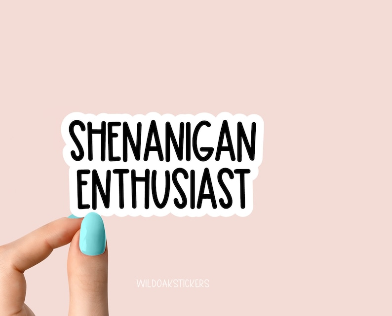 K&ouml;nnte beinhalten: Wei&szlig;er Aufkleber mit schwarzem Text, der "Shenanigan Enthusiast" lautet. Eine Hand h&auml;lt den Aufkleber.