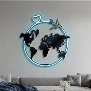 May include: A black metal world map wall art with a blue neon light outline. A small airplane and a heart shape are above the map. The art is mounted on a gray wall above a gray sofa with a blue blanket and a wooden coffee table.