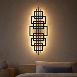 May include: A black wall-mounted light fixture featuring a vertical arrangement of interconnected rectangular frames. The geometric structure is backlit, casting a warm, soft glow against the wall surface behind the metal frame design.