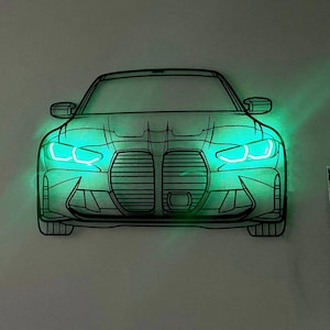 Pode incluir: Uma peça de arte de parede em metal preto com o formato da vista frontal de um carro, apresentando um design de grade de rim duplo. A peça é iluminada por trás com luzes LED verde brilhante que destacam os faróis e o contorno do carro.