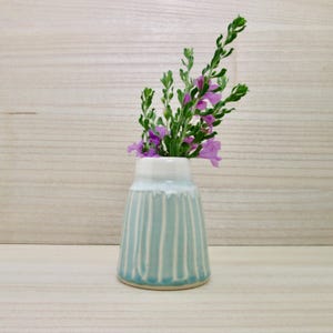 May include: A small ceramic vase with a light blue glaze and vertical ribbed texture, featuring a white drip pattern around the rim. The vase contains a sprig of green foliage with small purple flowers extending from the narrow opening.