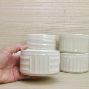 May include: A set of three off-white ceramic ramekins with a vertical ribbed texture on the exterior. Each small, round bowl features a glossy finish and a consistent cylindrical shape, designed for individual servings of food or small items.