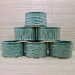 May include: A set of six small, cylindrical ceramic bowls with a light blue-green glaze. The bowls are stacked in a pyramid formation, featuring varied textured patterns including vertical lines and horizontal wavy indentations on the exterior.