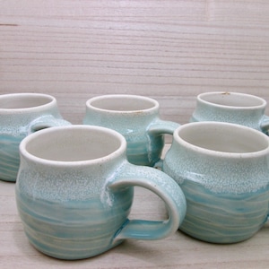 May include: A set of five ceramic mugs featuring a rounded shape and a handle. Each mug has a light blue base with horizontal ridges and a white, speckled glaze covering the upper portion and the interior rim of the vessel.