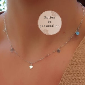Puede incluir: Un collar de cadena de plata con cuatro pequeños colgantes en forma de corazón. Dos de los colgantes son plateados y dos son azul claro. El collar lo lleva una persona con la piel bronceada. El texto "Option to personalise" está escrito en un círculo en la imagen.