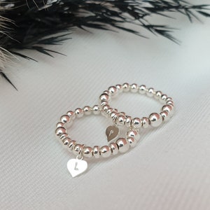 May include: Two silver beaded rings, each with a heart-shaped charm. One charm has the letter "L" and the other has the letter "P". The rings are displayed on a white surface, with dark green and brown foliage in the background.