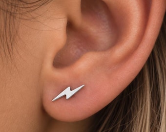 Puede incluir: Un pequeño pendiente de botón de color plateado con forma de rayo. El pendiente presenta un acabado metálico liso y está colocado en el lóbulo inferior de una oreja. El diseño es sencillo, con bordes afilados y angulares que forman el rayo.