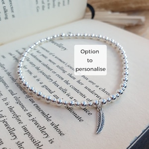 Può includere: Un braccialetto d'argento con perline e un ciondolo a forma di piuma. Il braccialetto è su un libro con il testo "Option to personalise" scritto su un quadrato bianco.