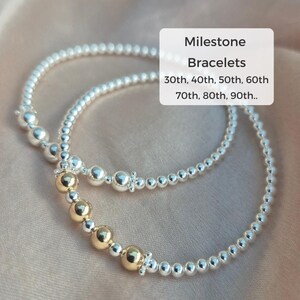 Op de afbeelding: Twee zilveren kralen armbanden met gouden kralen. De armbanden zijn op elkaar gestapeld. De tekst "Milestone Bracelets" staat boven de armbanden. De tekst "30th, 40th, 50th, 60th 70th, 80th, 90th.." staat onder de armbanden.