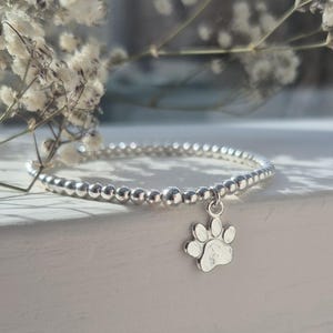 May include: A silver beaded bracelet with a paw print charm. The bracelet is made of small, round beads and features a dangling charm shaped like a dog or cat paw. The bracelet is displayed on a white surface.