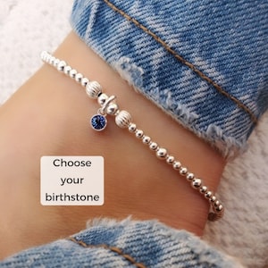 Puede incluir: Una pulsera de cuentas plateada con un pequeño dije de piedra preciosa azul. La pulsera se muestra en una muñeca con jeans en el fondo. El texto en la imagen dice "Elige tu piedra de nacimiento."