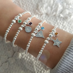Può includere: Quattro braccialetti d'argento con perline, ciondoli e pietre preziose colorate. I ciondoli includono un disco con la lettera "E", un cuore con la lettera "S" e una stella con la lettera "A". Le pietre sono verdi, viola, blu e arancioni.