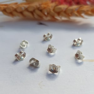 May include: Eight silver earring backs, also known as butterfly backs, are arranged on a white surface. The backs are designed to secure earrings to the earlobe.