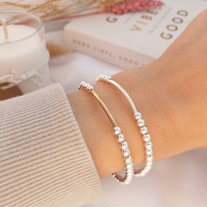 May include: Two bracelets on a wrist. One bracelet is silver with small beads and the other is a gold bar bracelet.