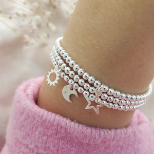 Op de afbeelding: Een zilveren armband met drie rijen kralen en drie bedels: een zon, een halve maan en een ster.