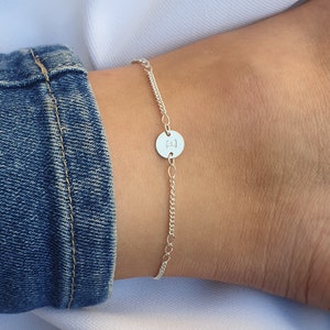 Puede incluir: Un brazalete de tobillo plateado con un pequeño dije redondo con la letra "H".
