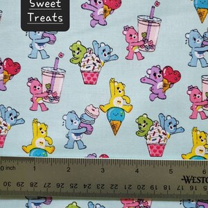May include: A light blue fabric with a repeating pattern of cartoon bears holding various sweet treats. The bears are in different colors and are holding ice cream cones, cupcakes, and bubble tea. The fabric has the text "Sweet Treats" printed on it.