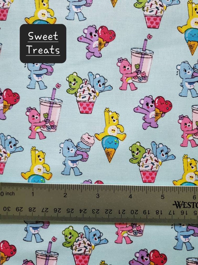 May include: A light blue fabric with a repeating pattern of cartoon bears holding various sweet treats. The bears are in different colors and are holding ice cream cones, cupcakes, and bubble tea. The fabric has the text "Sweet Treats" printed on it.