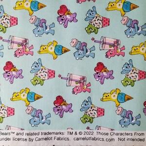 May include: A light blue fabric with a repeating pattern of Care Bears characters. The bears are holding ice cream cones and cups of bubble tea. The text "Care Bears&trade; and related trademarks: TM & &copy; 2022 Those Characters From Cleveland, L. Used under license by Camelot Fabrics. camelotfabrics.com Not intended for children." is printed at the bottom of the fabric.