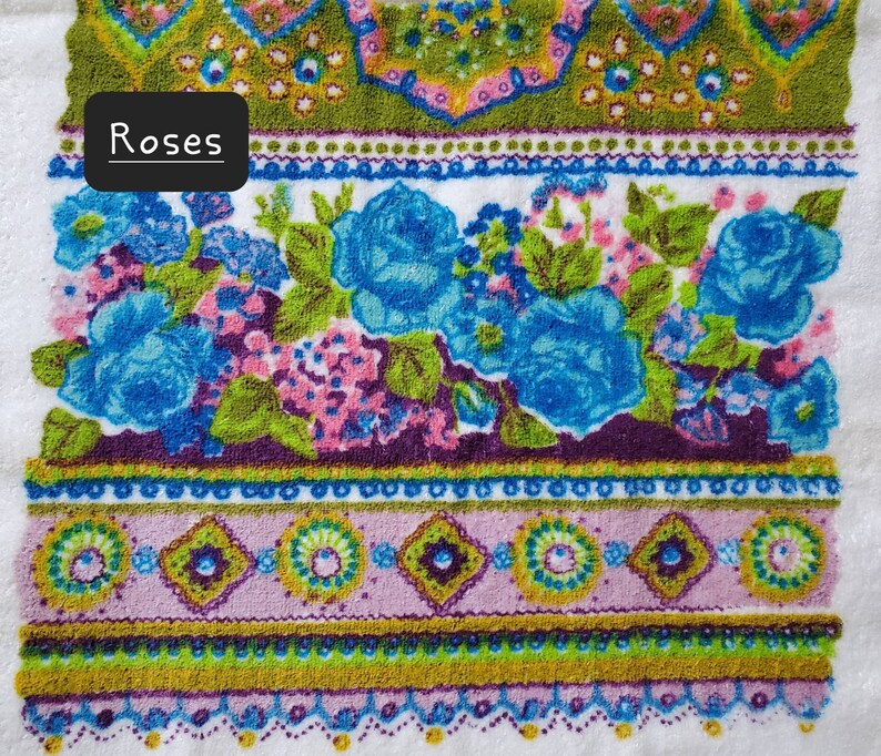 May include: A white towel with a colourful floral pattern. The pattern features blue roses, green leaves, and pink flowers. The towel has a border of green, purple, and pink.