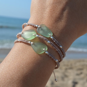 May include: Three layered bracelets featuring smooth, translucent light green sea glass pieces set on thin, braided beige cord. Each bracelet is accented with small, round silver-toned metal beads spaced evenly along the cord, creating a delicate design.