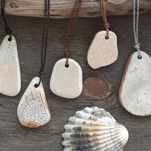 May include: Five ceramic pendants with different textures and shapes, hanging on cords against a wooden background. The pendants are white with brown accents and have a hole at the top for stringing.