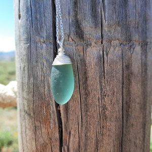 Op de afbeelding: Een zilveren ketting met een lichtblauwe zeeglas hanger gewikkeld in zilverdraad.