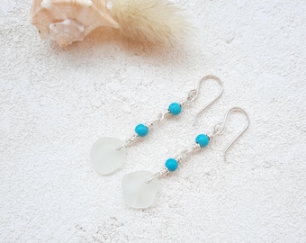 May include: Dangle earrings with sea glass pendants and turquoise beads. The earrings have silver-coloured hooks. They are displayed on a textured white surface, with a seashell in the background. The earrings are a beach-themed accessory.