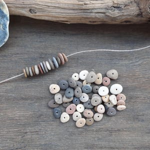 May include: A collection of natural, drilled stone beads in various colors, including gray, white, and pink, are displayed on a wooden surface. Some beads are strung on a white cord, while others are loose, suggesting crafting or jewelry making.