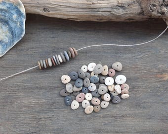 May include: A collection of natural, drilled stone beads in various colors, including gray, white, and pink, are displayed on a wooden surface. Some beads are strung on a white cord, while others are loose, suggesting crafting or jewelry making.