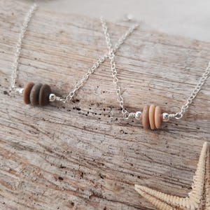 May include: Two delicate silver chain necklaces with a single charm each. One charm is a stack of three brown and beige sea glass stones. The other charm is a stack of three beige sea glass stones.