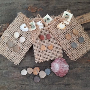 May include: Three burlap squares with different colored stones and shells attached. The stones are various shades of brown, gray, and white. The shells are white and beige. The squares are arranged in a triangle shape on a wooden surface.