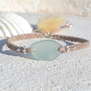 May include: A handcrafted bracelet with a pale blue, oval sea glass centrepiece. The bracelet is made with a woven, tan-coloured cord and small silver beads. A seashell and a feather are in the background.