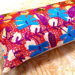 May include: A colorful embroidered pillow with a floral pattern in shades of red, purple, blue, and yellow. The pillow is made of a soft, white fabric and has a rectangular shape.