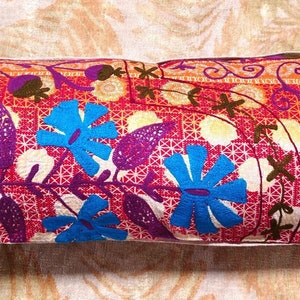 May include: A long, rectangular pillow with a colorful floral pattern. The pillow is made of a soft, textured fabric and features a variety of blue, purple, and orange flowers on a red background.