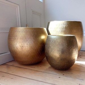 May include: Three rounded, metallic gold planters of varying sizes, each featuring a subtle, repeating geometric pattern etched into the surface. The containers have a wide, open top and a curved, bulbous body, reflecting light across their textured finish.