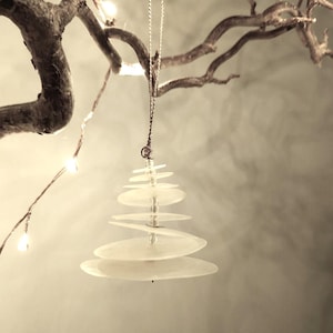 May include: A white, layered, circular ornament shaped like a Christmas tree hanging from a branch.