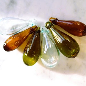 May include: A collection of six glass teardrop ornaments in various shades of green, brown, and clear. The ornaments are arranged in a fan shape on a white marble surface.